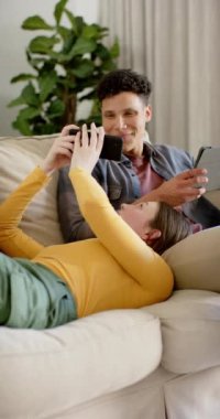 Kanepede oturan mutlu çiftin tablet ve akıllı telefonu yavaş çekimde kullandığının dikey videosu. İlişki, birliktelik, iletişim, yaşam tarzı ve aile hayatı, değişmedi..