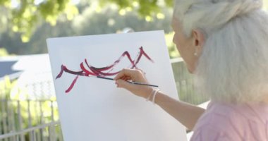 Evde balkonda resim yapan son sınıf melez mutlu kadın, ağır çekimde. Ev hayatı, emeklilik, sanat, doğa ve yaşam tarzı, değiştirilmemiş.