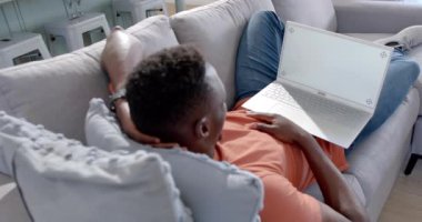 Mutlu Afro-Amerikalı adam yalan söylüyor ve güneşli oturma odasında bilgisayarına video çekiyor, ağır çekimde. Ev, ev hayatı, yaşam tarzı, refah, iletişim, teknoloji, değişmemiş..