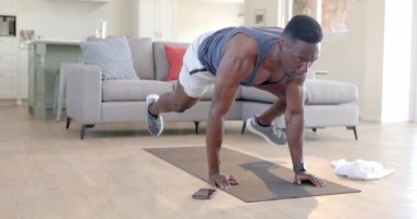 Odaklanmış Afro-Amerikan bir adam oturma odasında akıllı telefonuyla dağcıları beceriyor, ağır çekimde. Ev, egzersiz, fitness ve sağlıklı yaşam tarzı, iletişim, değişmemiş.
