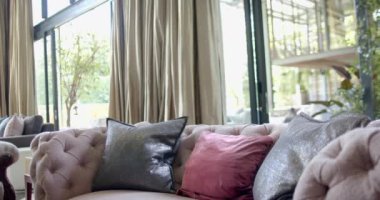 Yastıklı bej kanepe, altın perdeler ve güneşli oturma odasında kocaman pencereler, yavaş çekim. İç tasarım, ev ve ev hayatı.