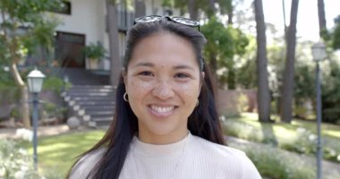 Güneş gözlüklü mutlu Asyalı kadın güneşli bahçede ayakta ve gülümsüyor, ağır çekimde. Yaz, tatil, doğa, refah, değişmemiş.