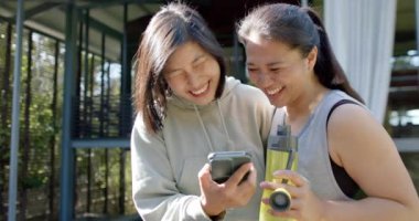 Su şişesi ve akıllı telefonlu mutlu Asyalı bayan arkadaşlar terasta gülüyorlar, ağır çekimde. Sağlıklı yaşam tarzı, zindelik ve refah, arkadaşlık, iletişim, değişmemiş.