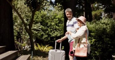 Güneşli ve açık havada bir eve bavullarıyla yürüyen mutlu yaşlı çift. Emeklilik, dinlenme, seyahat, inşaat ve doğa konsepti, değiştirilmemiş..