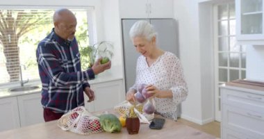 Mutfaktaki alışveriş çantalarını boşaltan son sınıf melez çift, yavaş çekimde. Ev hayatı, emeklilik, yemek ve yaşam tarzı, değişmemiş..