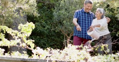 Mutlu çift ırklı yaşlı çiftler güneşli bahçede çiçekleri suluyor. Emeklilik, dinlenme, boş zaman, birliktelik ve doğa konsepti, değiştirilmemiş..