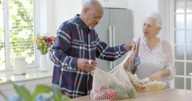 Mutfaktaki alışveriş çantalarını boşaltan son sınıf melez çift, yavaş çekimde. Ev hayatı, emeklilik, yemek ve yaşam tarzı, değişmemiş..