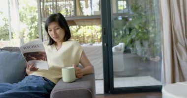 Mutlu Asyalı kadın elinde fincanla kanepede oturuyor ve güneşli oturma odasında kitap okuyor, ağır çekimde. Yaşam tarzı, rahatlama, boş zaman, refah ve ev hayatı, değişmemiş..
