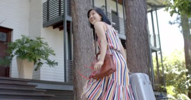 Güneşli bir günde valizli mutlu Asyalı kadın evinin merdivenlerinde yürüyor, ağır çekimde. Yaz, eve dönüş, seyahat, değişmeden.