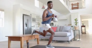 Odaklanmış Afro-Amerikan bir adam güneşli oturma odasında kettlebell ile egzersiz yapıyor, ağır çekimde. Ev, egzersiz, fitness ve sağlıklı yaşam tarzı, değişmemiş..