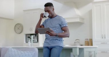 Mutlu Afro-Amerikan adam kahve içiyor ve güneşli mutfakta tablet kullanıyor, ağır çekimde. Ev, ev hayatı, yaşam tarzı, yemek, rahatlama, iletişim, değişmemiş.