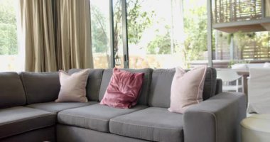 Yastıklı kahverengi kanepe, altın perdeler ve güneşli oturma odasında kocaman pencereler, yavaş çekim. İç tasarım, ev ve ev hayatı.