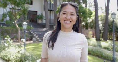 Güneş gözlüklü mutlu Asyalı kadın güneşli bahçede ayakta ve gülümsüyor, ağır çekimde. Yaz, tatil, doğa, refah, değişmemiş.