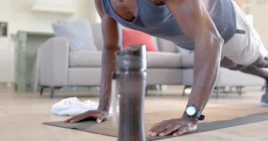 Odaklanmış Afro-Amerikan bir adam güneşli oturma odasında su şişesinin yanında şınav çekiyor, ağır çekimde. Ev, egzersiz, fitness ve sağlıklı yaşam tarzı, değişmemiş..