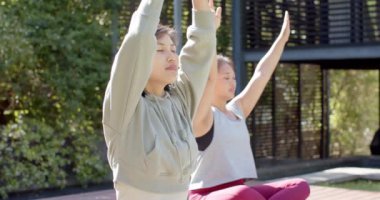 Odaklanmış Asyalı bayan arkadaşlar güneşli terasta yoga meditasyonu yapıyorlar, yavaş çekim. Sağlıklı yaşam tarzı, zindelik ve refah, arkadaşlık, değişmemiş.