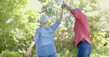 Mutlu son sınıf melez çift evde güneşli bahçede dans ediyor, ağır çekimde. Ev hayatı, emeklilik, doğa ve yaşam tarzı, değişmemiş..
