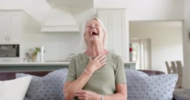 Kafkasyalı son sınıf öğrencisi mutlu bir kadın güneşli oturma odasında el sallıyor, ağır çekimde. Emeklilik, ev hayatı ve yaşlılar yaşam tarzı, iletişim, teknoloji, değişmemiş.