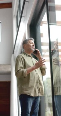 Evde akıllı telefondan konuşan yaşlı, çift ırklı bir adamın dikey videosu, yavaş çekim. Boş zaman, iletişim, teknoloji, ev hayatı ve yaşlı yaşam tarzı, değiştirilmemiş.
