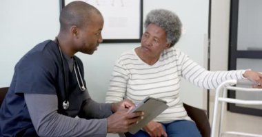 Afro-Amerikan erkek doktor tablet kullanıyor ve hastanede yaşlı kadınla konuşuyor, ağır çekimde. Tıp, sağlık, iletişim ve tıbbi hizmetler, değiştirilmemiş.