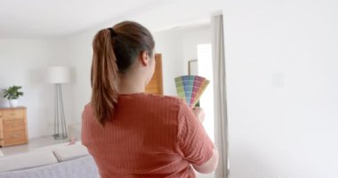 Düşünceli ve melez bir kadın güneşli oturma odasında renkli tabloya bakıyor, ağır çekimde. Ev dekorasyonu, iç tasarım, diy, ev hayatı ve yaşam tarzı, değiştirilmemiş.