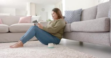 Mutlu ve çift ırklı kadın oturma odasında, tablet, kopyalama alanı ve ağır çekim kullanarak rahatlıyor. İletişim, teknoloji, rahatlama, ev hayatı ve yaşam tarzı, değiştirilmemiş.