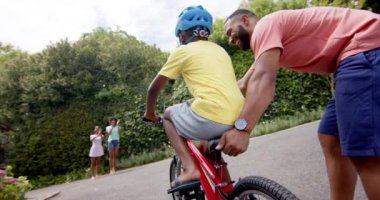 Afrikalı Amerikalı baba oğluna bisiklet sürmeyi öğretiyor, anne ve kız kardeş alkışlıyor, ağır çekimde. Çocukluk, babalık, aile, öğrenme, bisiklet, yaz, birliktelik ve yaşam tarzı, değişmemiş..