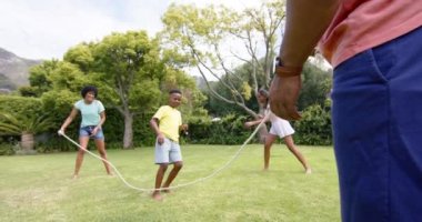 Mutlu Afro-Amerikan ebeveynler, oğlu ve kızı güneşli bahçede zıplıyorlar, ağır çekimde. Çocukluk, aile, yaz, boş zaman, eğlence, birliktelik ve yaşam tarzı, değişmemiş..