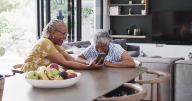 Kıdemli Afro-Amerikan bayan arkadaşları akıllı telefon kullanıyor ve yemek odasında gülüyorlar, ağır çekimde. İletişim, rahatlama, boş zaman, arkadaşlık, birliktelik ve kıdemli yaşam tarzı, değişmemiş..