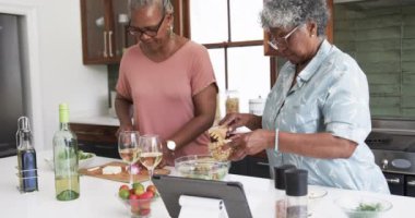 Odaklanmış kıdemli Afro-Amerikan bayan arkadaşları yemek pişiriyor ve tablet kullanıyor, kopyalama alanı, ağır çekim. İletişim, yemek, yemek, arkadaşlık, refah ve sağlıklı yaşlı yaşam tarzı, değişmemiş.