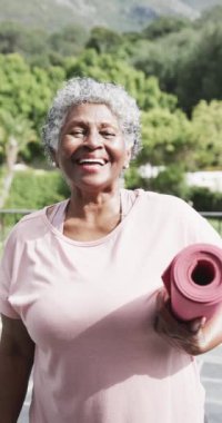 Bahçesinde yoga minderi olan mutlu bir Afrikalı Amerikalı kadının dikey video portresi, yavaş çekim. Yoga, fitness, refah ve sağlıklı yaşlı yaşam tarzı, değişmemiş.
