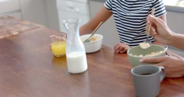 Çift ırklı anne ve kız kahvaltı gevreğini güneşli mutfakta yavaş çekimde yiyorlar. Annelik, çocukluk, birliktelik, yemek, yemek, sabah, ev hayatı ve yaşam tarzı, değişmemiş.