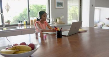 Kulaklıklı melez bir kız ilkokul dersini laptopta, kopyalama alanında, ağır çekimde alıyor. Eğitim, iletişim, okul, öğrenme, çocukluk, teknoloji ve ev hayatı, değiştirilmemiş.