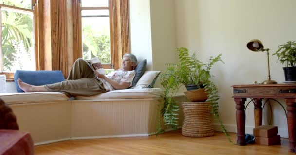 快乐的老女人坐在沙发上 在家里的窗口看书 慢动作 家庭生活 退休和生活方式 — 图库视频影像