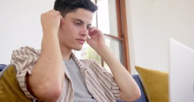 Kulaklık takan çift ırklı adam oturma odasında dizüstü bilgisayar kullanarak koltukta oturuyor, ağır çekimde. İletişim, teknoloji, ev hayatı ve yaşam tarzı, değişmedi..