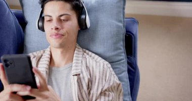 Kulaklıklı mutlu bir çift ırklı adam kanepeye uzanmış akıllı telefon, kopyalama alanı ve ağır çekim kullanıyor. İletişim, müzik, teknoloji, rahatlama, ev hayatı ve yaşam tarzı, değiştirilmemiş.