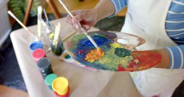 Üst düzey melez kadın önlük giyiyor ve palette renkleri karıştırıyor, ağır çekimde. Ev hayatı, sanat, emeklilik ve yaşam tarzı, değişmedi..