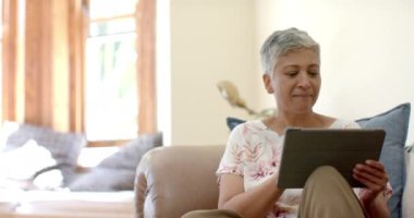 Mutlu son sınıf melez kadın koltukta oturup gülüyor ve tablet kullanıyor, ağır çekimde. Ev hayatı, teknoloji, emeklilik ve yaşam tarzı, değişmedi..