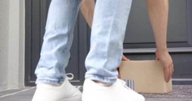 Çift ırklı adamın ön kapının dışındaki karton kutuda teslimat yapan alt kesimi, ağır çekim. Teslimat, çevrimiçi alışveriş, nakliye, posta servisi, paket, ev hayatı ve yaşam tarzı.