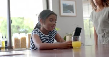 Güneşli mutfakta annesiyle birlikte akıllı telefon kullanan mutlu çift ırklı kız, yavaş çekim. Çocukluk, iletişim, teknoloji, sabah, ev hayatı ve yaşam tarzı, değişmemiş..