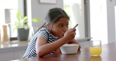 Çift ırklı bir kız kahvaltı gevreği yiyor güneşli bir mutfakta oturuyor, fotokopi odası, ağır çekim. Çocukluk, yemek, yemek, sabah, ev hayatı ve yaşam tarzı, değişmemiş..