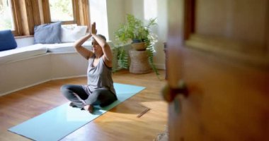 Evde yoga minderi üzerinde meditasyon yapan yaşlı çift ırklı kadın. Ev hayatı, yoga, yaşam tarzı, değişmemiş..