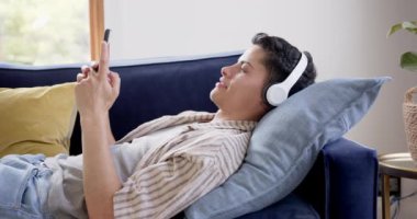 Kulaklıklı mutlu bir çift ırklı adam kanepeye uzanmış akıllı telefon kullanıyor, ağır çekimde. İletişim, müzik, teknoloji, rahatlama, ev hayatı ve yaşam tarzı, değiştirilmemiş.