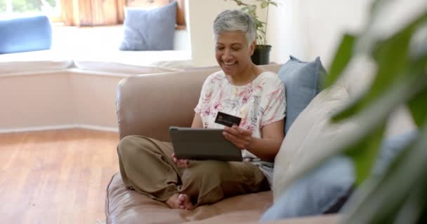 快乐的老女人坐在沙发上 手里拿着信用卡 在家里用平板电脑 慢动作 家庭生活 报酬和生活方式 — 图库视频影像