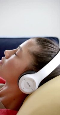 Kulaklık takmış, koltukta ağır çekimde gülümseyen melez bir genç kızın dikey videosu. Müzik, iletişim, ergenlik, rahatlama, ev hayatı ve yaşam tarzı, değişmemiş..