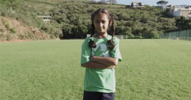 Çift ırklı bir kız yeşil bir geri dönüşüm tişörtü giyerek bir futbol sahasında güvenle duruyor. Güneşli bir ortamda, ağır çekimde takım çalışması ya da maça hazır..
