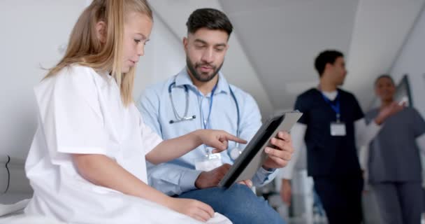 若いアジアの医師は コピースペースを持つ白人少女とタブレット上の医療情報をレビューします 彼らはスクリーンに焦点を当て 病院の設定で相談を提案しています — ストック動画