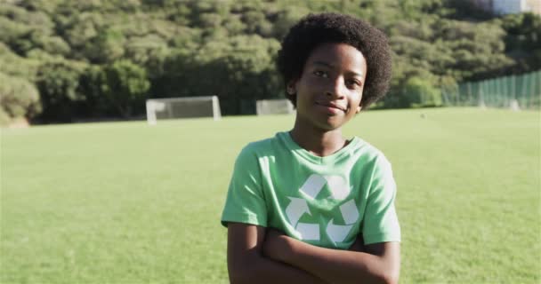 Biracialの少年は コピースペースを備えた緑色のリサイクルTシャツを着たサッカー場に自信を持って立っています 彼は屋外のシャツのリサイクルシンボルで環境意識を促進し スローモーション — ストック動画