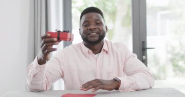 Afrikalı Amerikalı bir adam video görüşmesinde evde bir hediye sunar. Sıcak bir şekilde gülümsüyor, neşeli ve samimi bir atmosfer yaratıyor, ağır çekimde..