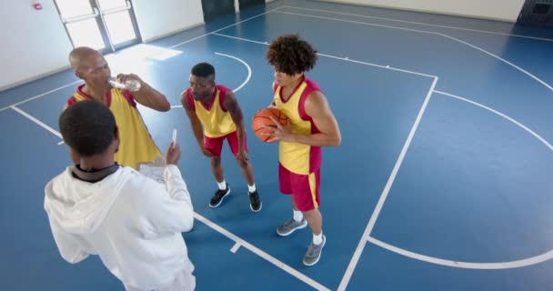 さまざまなバスケットボールチームが法廷で戦略を議論しています プレーヤーは 屋内バスケットボールゲームでタイムアウト中にコーチのプレイブックに焦点を当てています スローモーション — ストック動画