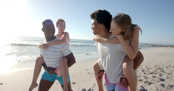 多様な家族が日当たりの良いビーチを満喫しています 笑顔の両親は 海で彼らの楽しい子供たちにピギーバック乗車を与え ゆっくりとした動き — ストック動画